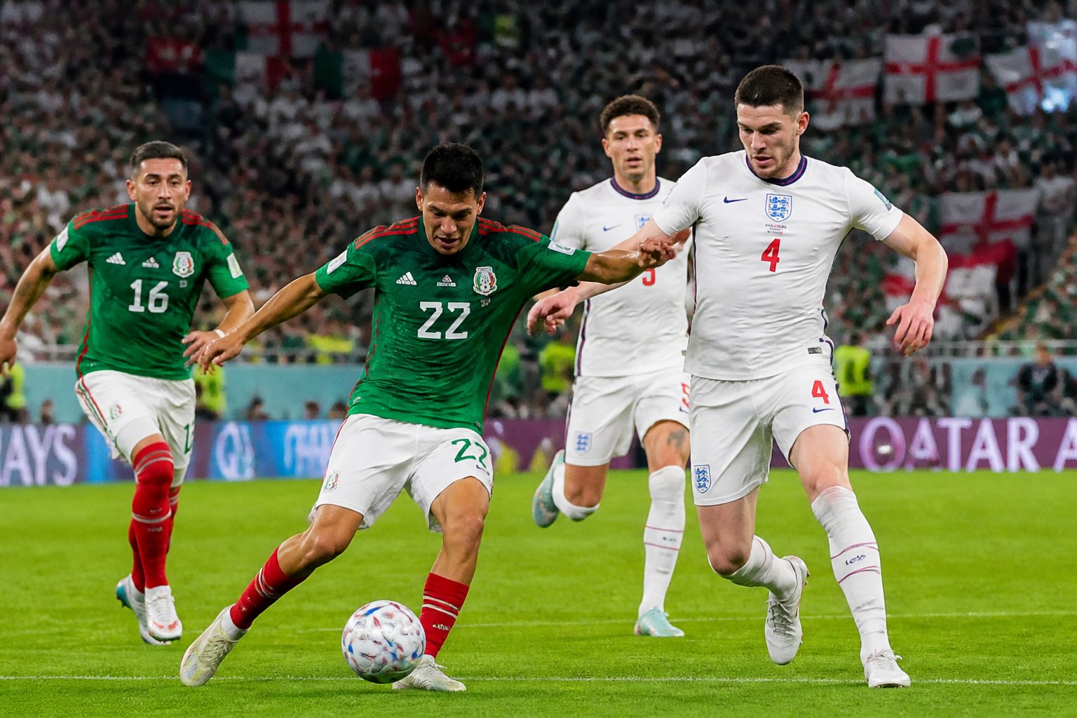 Jugadores de México e Inglaterra compitiendo por el balón en un partido de fútbol con un estadio lleno de aficionados al fondo.